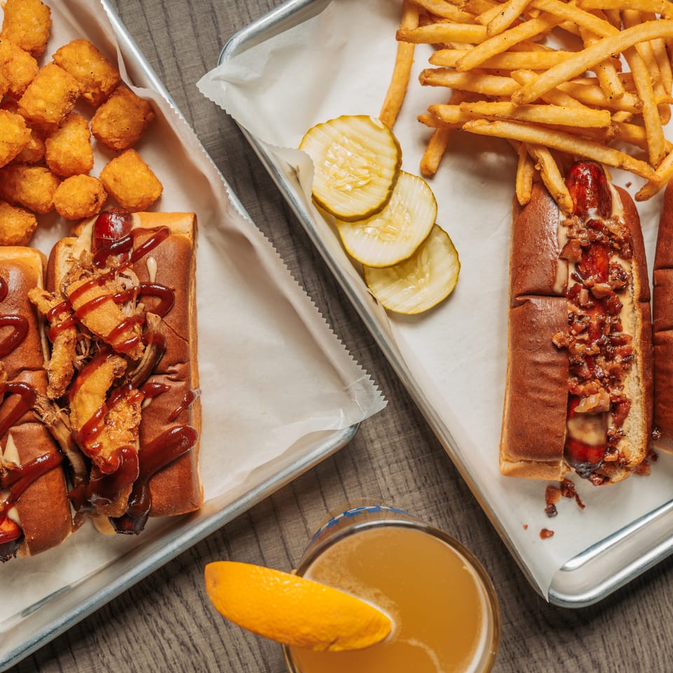 Two trays of hot dogs, one with tater tots and the other with fries, plus a drink.